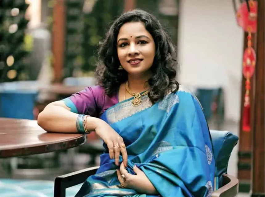 Dr. Rajitha Vanga in a blue silk saree, smiling, resting her arm on a table